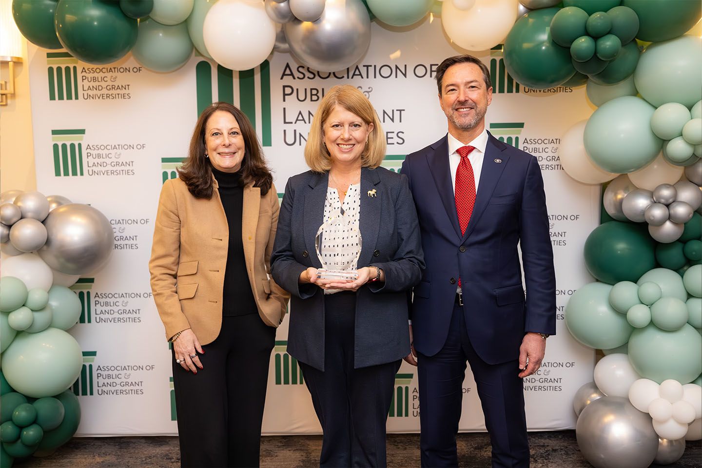 Marisa Pagnattaro, Stacy Jones, and Chris King with crystal award in front of APLU photo backdrop with a green and silver balloon arch