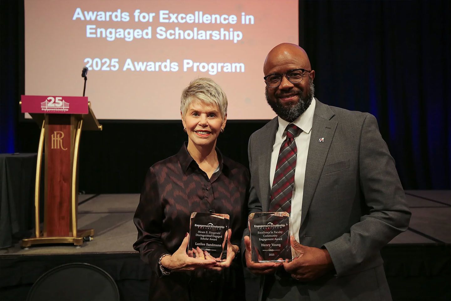two UGA faculty hold awards received at the ESC conference, the slideshow in the background reads "Awards for Excellence in Engaged Scholarship: 2025 Awards Program"